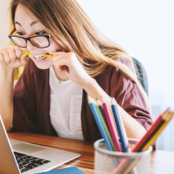 woman biting pencil while sitting on chair in front of computer during daytime