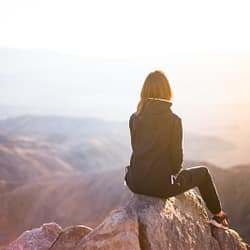 person sitting on top of gray rock overlooking mountain during daytime